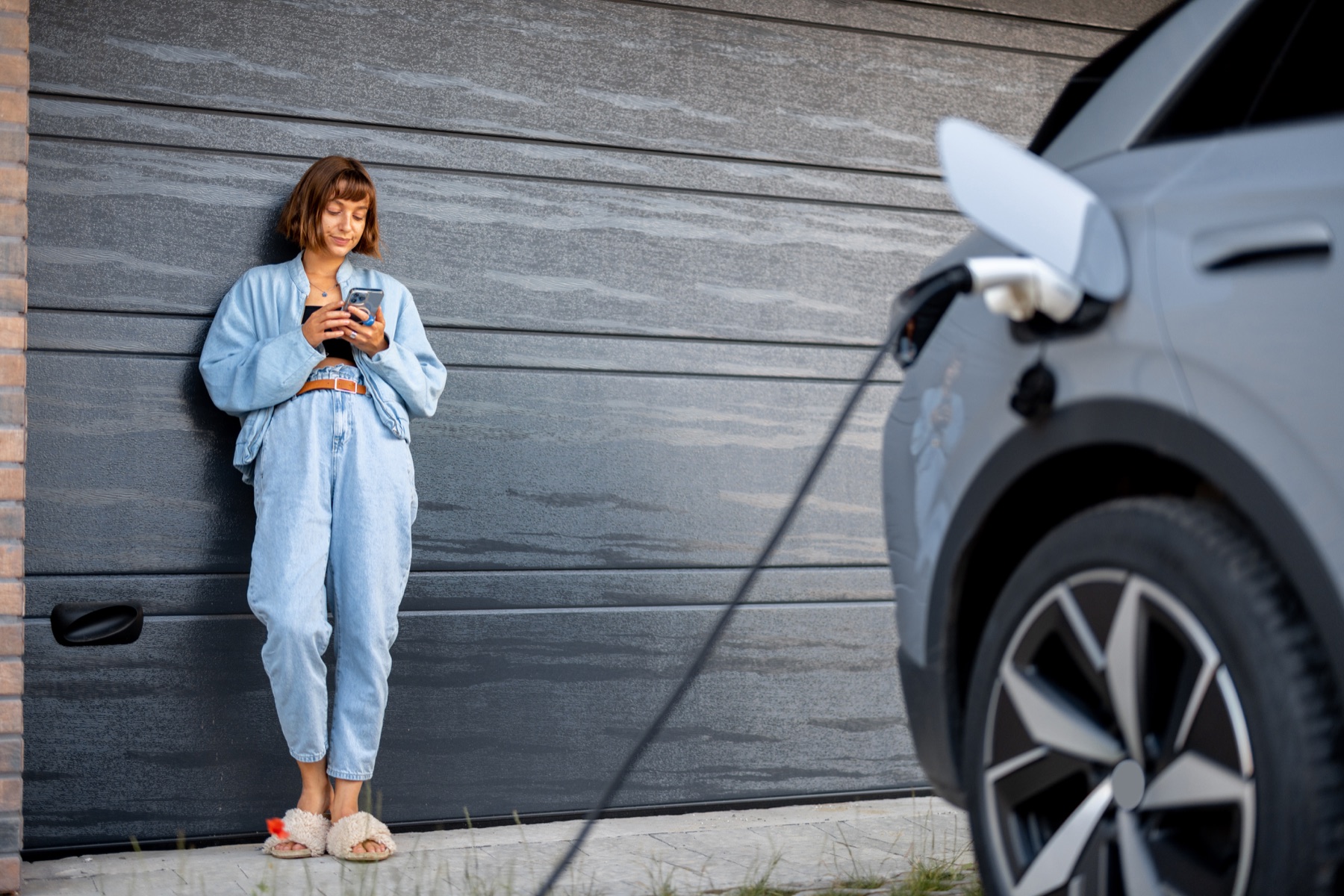 Mujer esperando con el celular mientras su auto se carga frente al portón del garaje